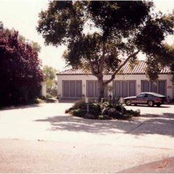 A large house with closed shutters and a parked car in front, surrounded by trees and shrubs on a sunny day.