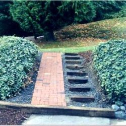 A brick ramp and uneven steps are surrounded by bushes with a grassy slope and trees in the background.